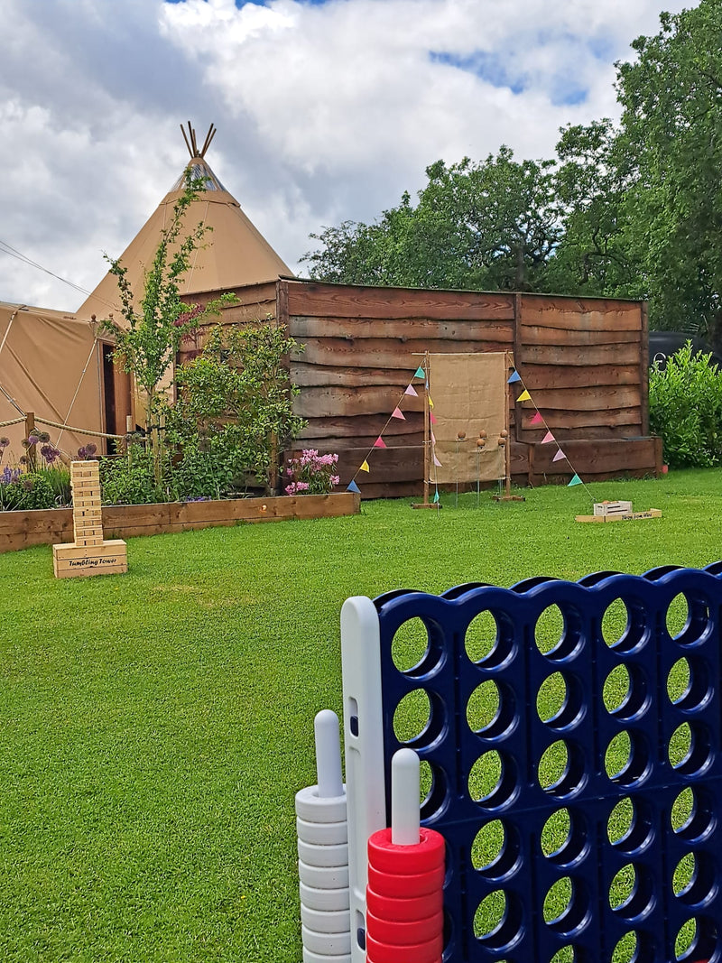 Outdoor scene at a wedding venue with a large tan tipi tent, coconut shy game on a grassed lawn, a blue 4 in a row game and giant tumbling towers in the foreground. The Tipis at Riley Green, Preston